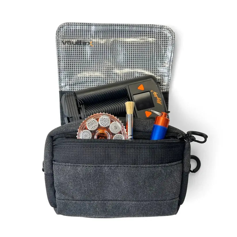 A dark gray Vaultlax fabric pouch shown open on a white background, containing various small tools and accessories including a black Mighty Plus Medic device, a round copper-colored dosing capsule magazine, a small brush, and a blue-and-orange dosing caddy. The pouch has a front zip pocket and a mesh-lined interior.