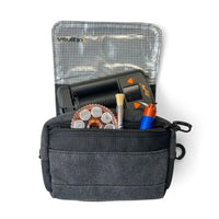 A dark gray Vaultlax fabric pouch shown open on a white background, containing various small tools and accessories including a black Mighty Plus Medic device, a round copper-colored dosing capsule magazine, a small brush, and a blue-and-orange dosing caddy. The pouch has a front zip pocket and a mesh-lined interior.