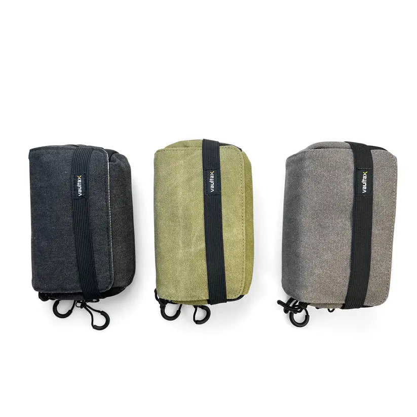 Three small rectangular fabric Vaultlax pouches or cases shown side by side on a white background, in dark charcoal gray, olive green, and heather gray, each with a black vertical strap and small white brand tag.